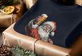A folded sweater with a print of Santa Claus holding a beer, lies on a wrapped gift. Surrounding pine cones and greenery add a festive touch.