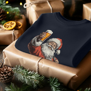 A folded sweater with a print of Santa Claus holding a beer, lies on a wrapped gift. Surrounding pine cones and greenery add a festive touch.