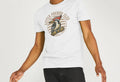 Man in a white "Little Pecker Club" T-shirt featuring a colourful woodpecker graphic. Wearing black jeans, he's mid-motion, smiling energetically.
