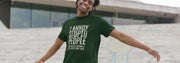 Young man smiling with arms wide open, wearing a green funny slogan T-shirt with humorous text. Steps and a modern building are in the background, creating a casual, playful tone.