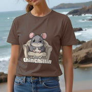 Woman at the beach wears a brown Graphic T-shirt with a cartoon chinchilla on a chair and text