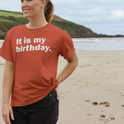 A smiling woman on a beach wearing a rust-colored Relaxed Fit Birthday T-shirt with
