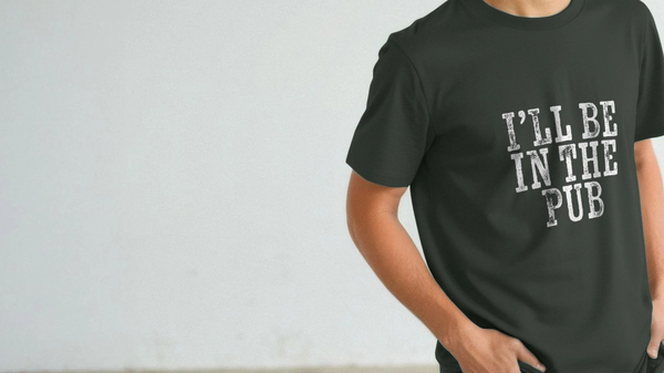A person wearing a black funny slogan T-shirt with the playful text, "I'LL BE IN THE PUB," printed in white. The background is a plain, light gray wall.