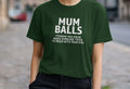 A woman on a cobblestone street wears a green t-shirt with bold white text: "Mum Balls. Things You Grow When Someone Tries to Mess With Your Kid." She appears content.