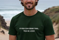 Smiling man on a beach wearing a dark green T-shirt with the joke text, "If you can read this... pass me a beer." The mood is light-hearted and casual.