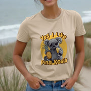 A woman stands on the beach, smiling, wearing a beige Women's Animal t-shirt with a cute koala graphic holding a pineapple and drink. The text reads 
