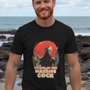 A smiling man stands on a rocky beach, wearing a black funny offensive T-shirt featuring an illustration of a giant rooster in a city with text reading 