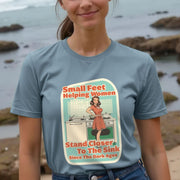 Smiling woman wearing a blue funny T-shirt with a retro-style graphic. The graphic depicts a woman by a stove, with text about women standing closer to the sink. Ocean and rocks in the background.