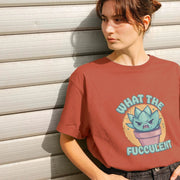 A Woman stands against a white, horizontal-paneled wall, wearing a red Graphic T-shirt with a cartoon Succulent and the text 