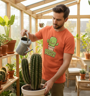Man watering a cactus in a greenhouse wearing an orange t-shirt with a cactus graphic.